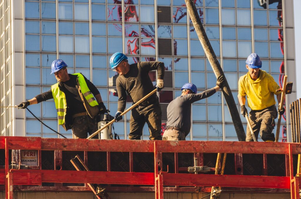 Vier bouwvakkers met blauwe helmen storten beton op een bouwplaats voor een glazen kantoortoren