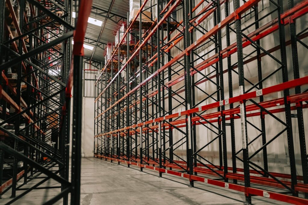 Hoge palletstellingen met rode liggers in een grotendeels lege warehousehal met daglicht door het dak