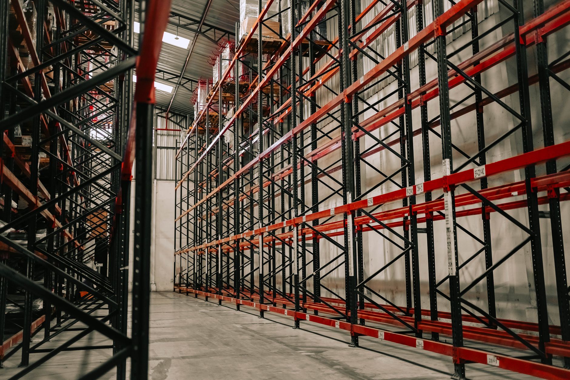 Hoge palletstellingen met rode liggers in een grotendeels lege warehousehal met daglicht door het dak