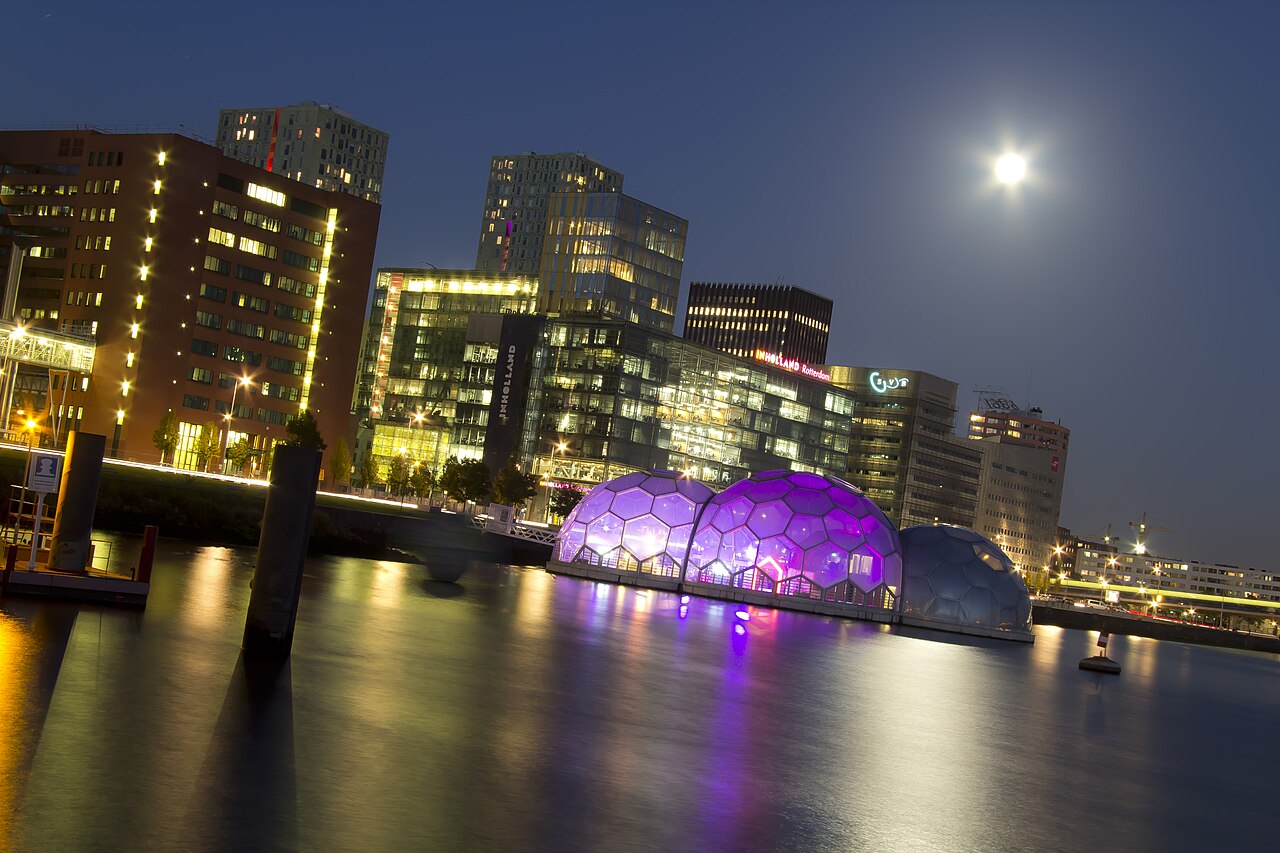 Verlichte kantoortorens aan de Rijnhaven op de Kop van Zuid in Rotterdam bij avond, met de drijvende paviljoens op het water